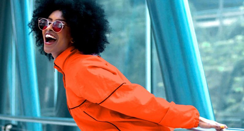 Power für den Tag Eine Frau mit Afrofrisur und orange-farbener Jacke und Sonnenbrille, lacht fröhlich.